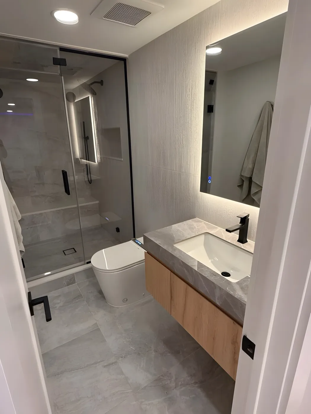 Floating wood vanity with a gray top, black faucet and backlit mirror in a basement bathroom