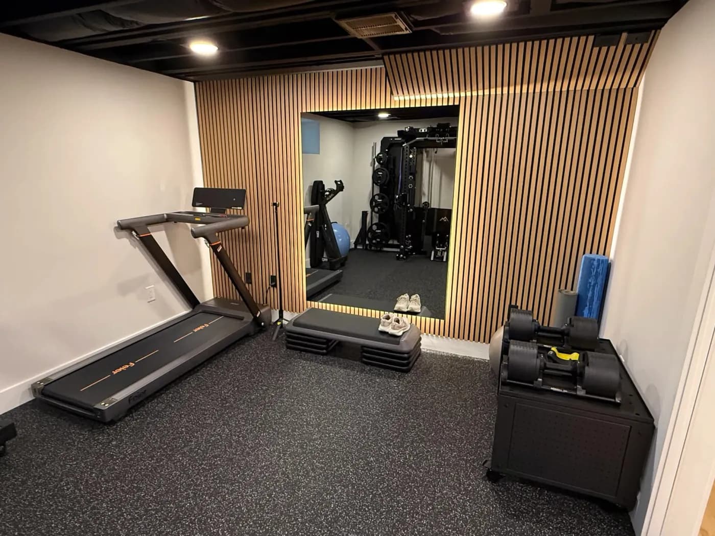 Basement home gym with rubber flooring, a wood slat wall, a large mirror and a black open ceiling