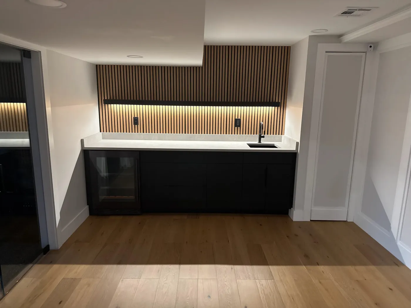 Basement wet bar with black cabinets, a drink fridge and a lit wood slat wall