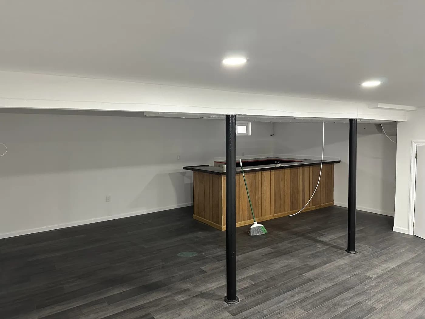 Rebuilt basement with gray wood-look plank flooring, black steel support columns, recessed lights and a wood-paneled bar