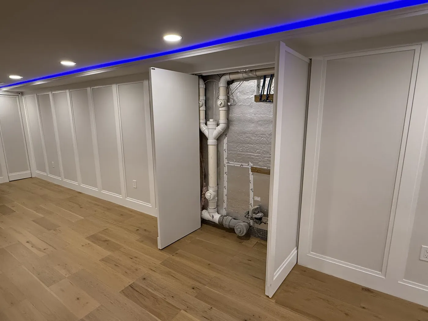 Paneled basement wall with two hidden doors open to show drain and water piping behind them