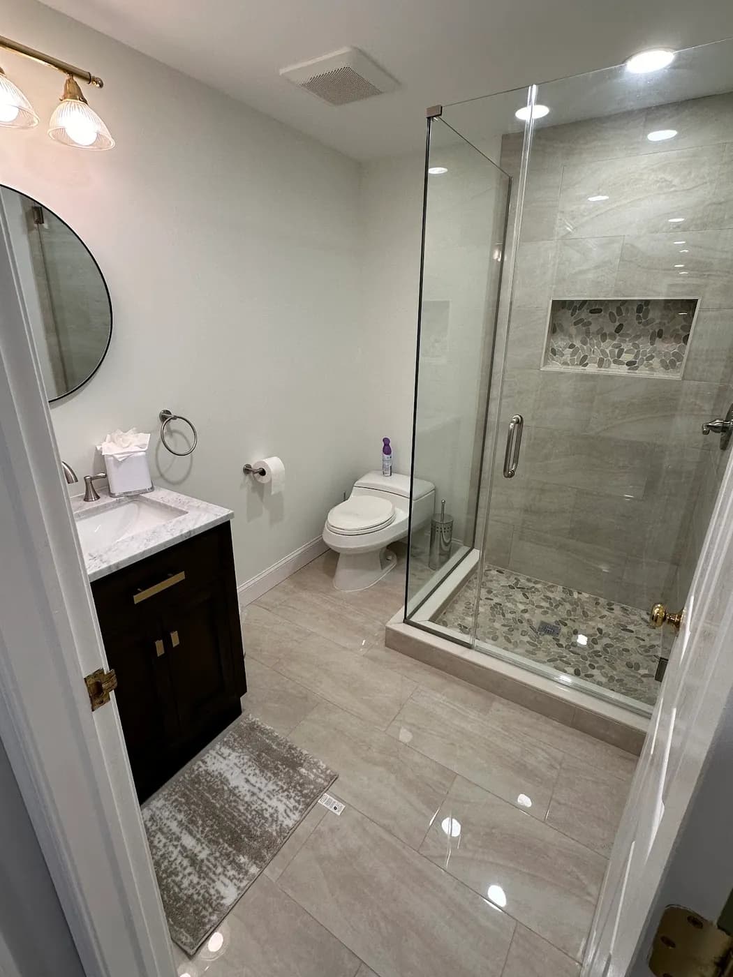 Bathroom with a frameless glass shower, a tiled shower niche, a dark wood vanity, a round mirror and large porcelain floor tile