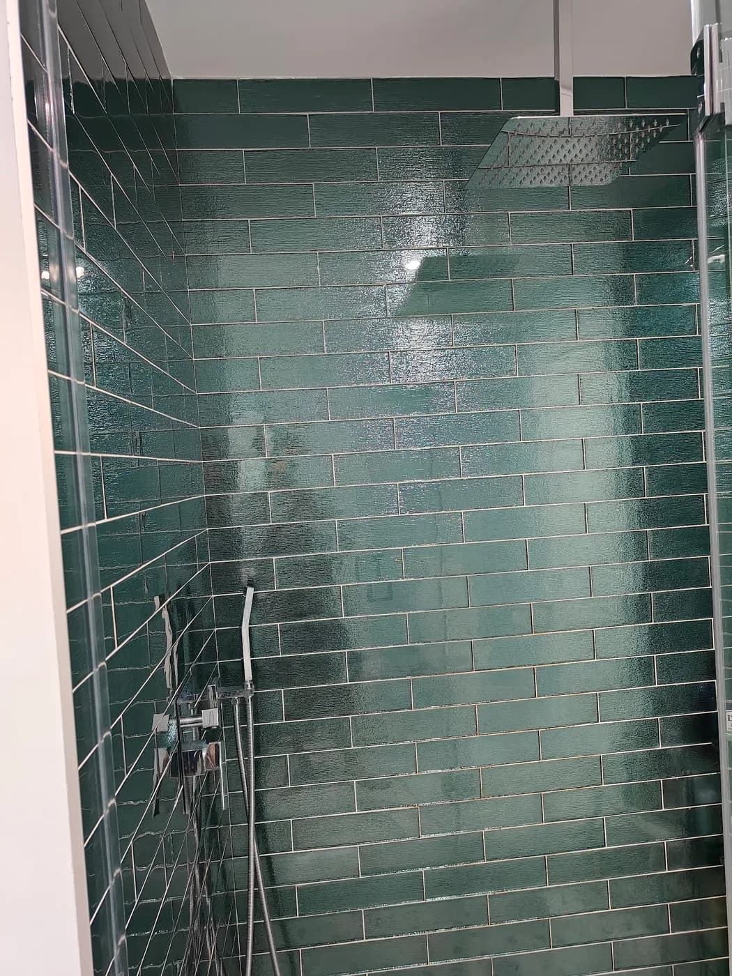 Shower lined with glossy green subway tile, with a ceiling rain shower head, handheld sprayer and glass enclosure