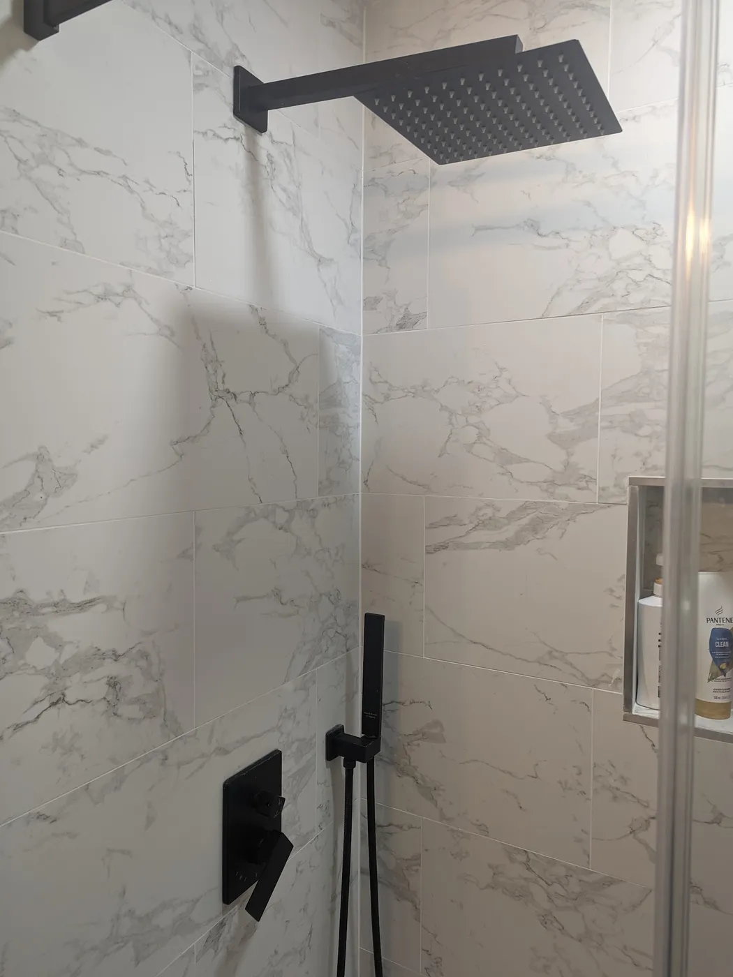 Walk-in shower with large marble-look wall tile, a matte black rain shower head, handheld sprayer and a recessed niche