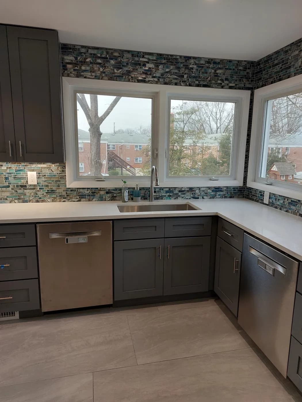Kitchen corner with a sink under a wide window, gray lower cabinets, light counters and large gray floor tile