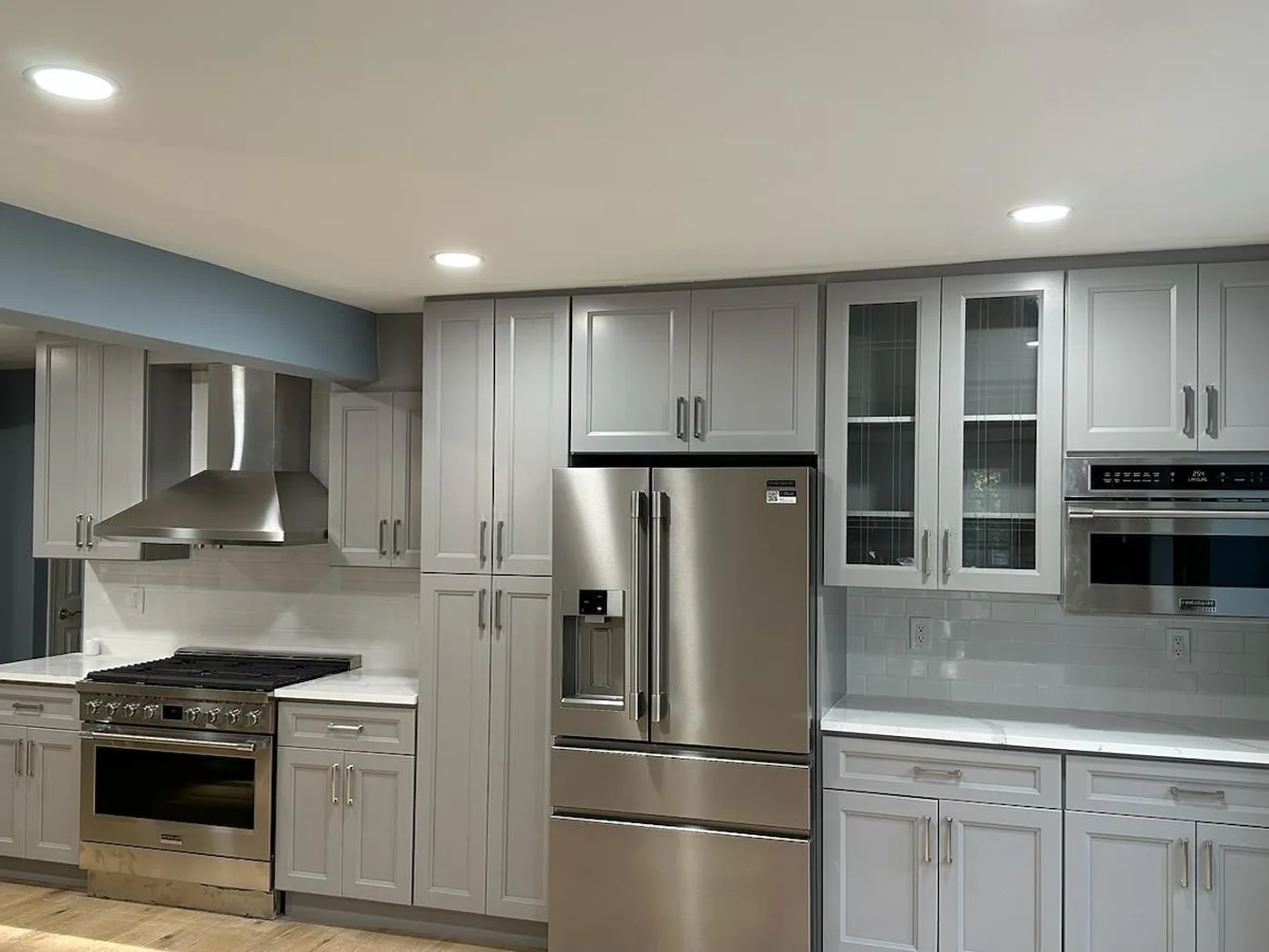 Light gray kitchen cabinets with glass-front uppers, a stainless refrigerator, range, hood and wall oven