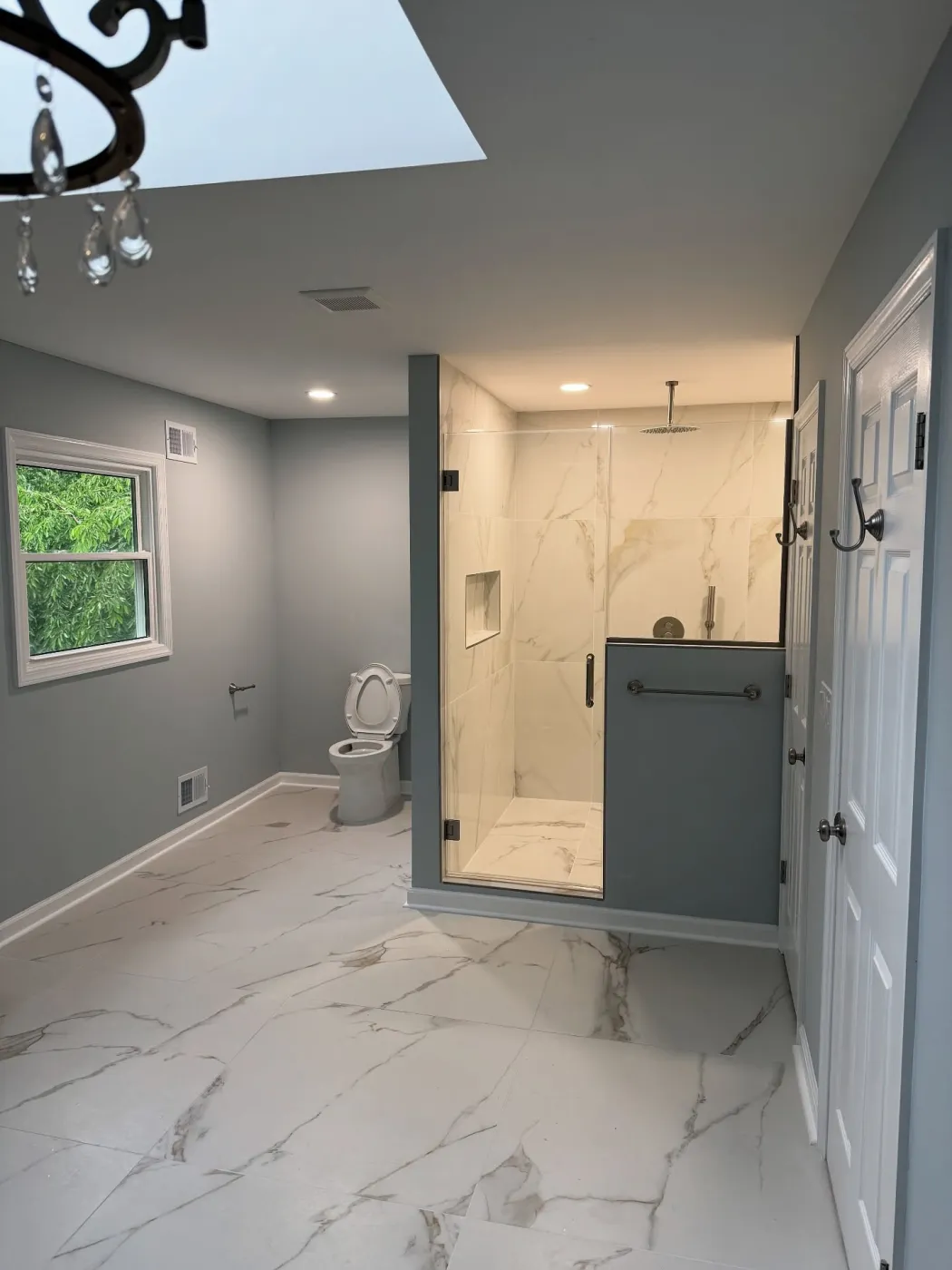 Bathroom with a skylight, gray-green walls, a white veined floor and a glass shower at the far end