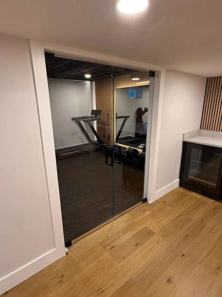 Frameless glass double doors open onto a basement gym with rubber floor, beside a wine fridge and white quartz counter.