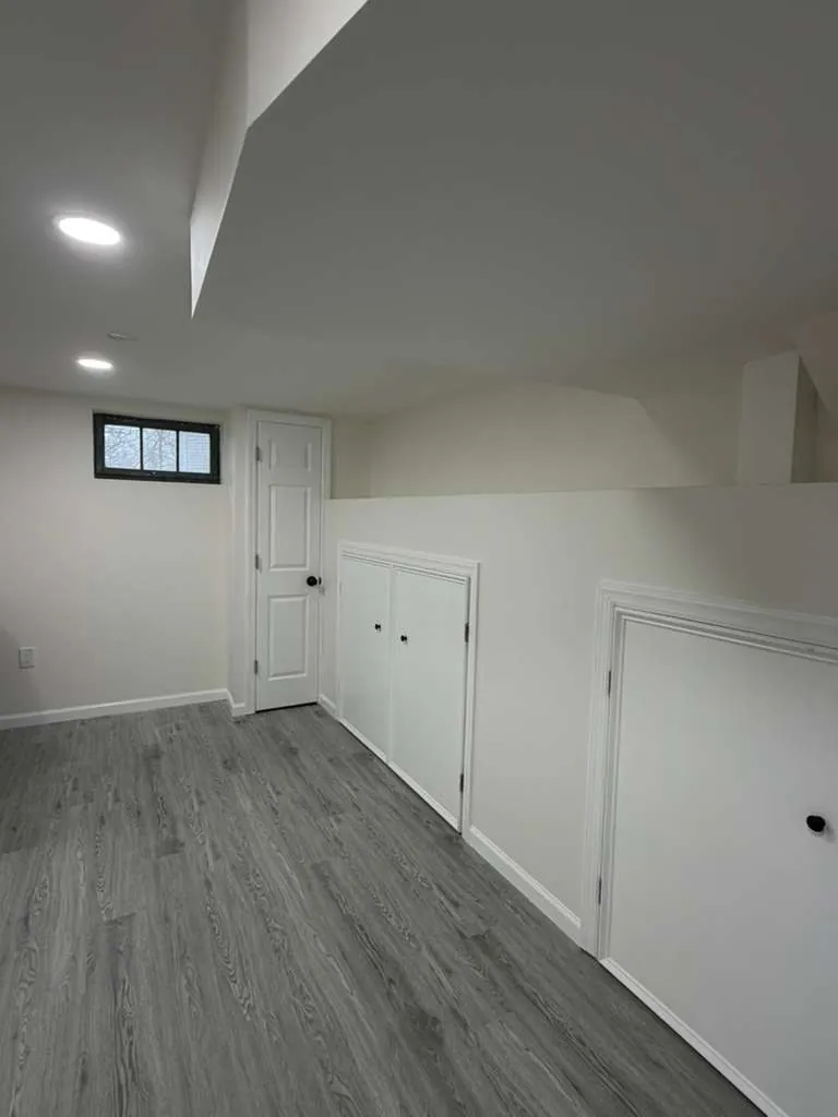 Basement room with white walls and trim, gray plank flooring, a small black-framed window and built-in cabinet doors.