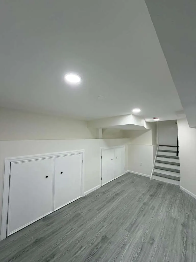 Finished basement with gray vinyl plank flooring, recessed lights and white built-in storage cabinet doors in the knee wall.