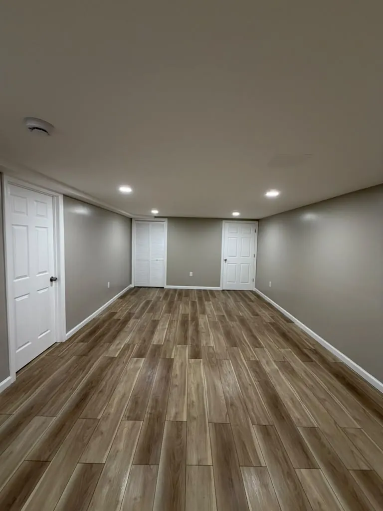 Long basement room with gray walls, wood-look plank floor, recessed lights, a louvered closet and white doors at the end.