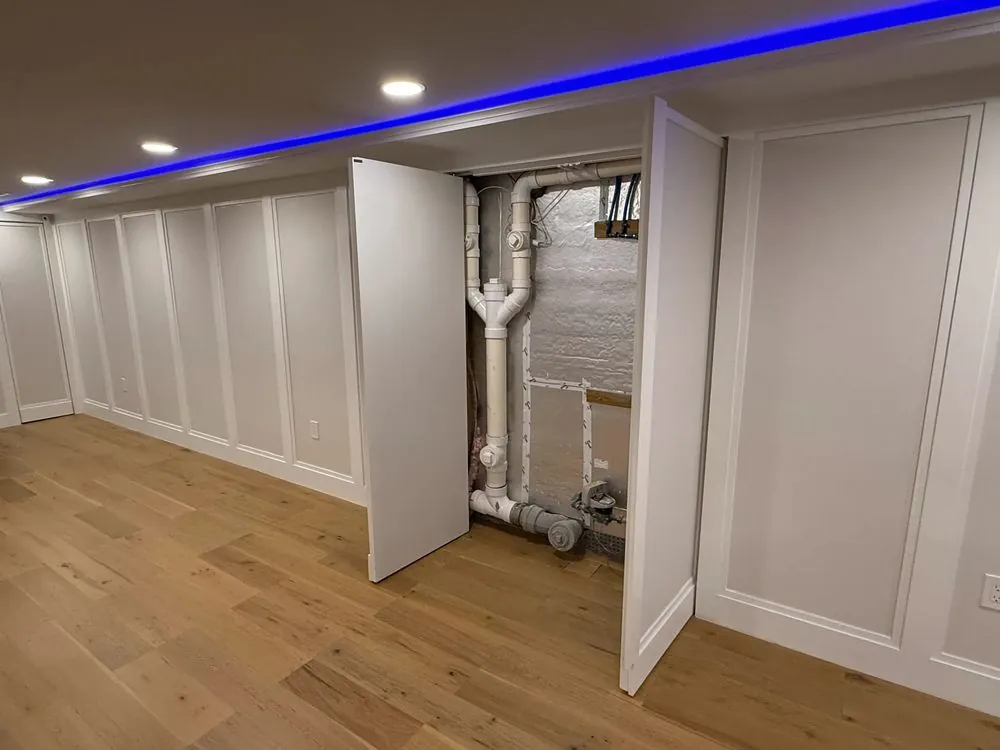 White paneled basement wall with two hinged panels opened to reveal hidden plumbing pipes and a water meter.