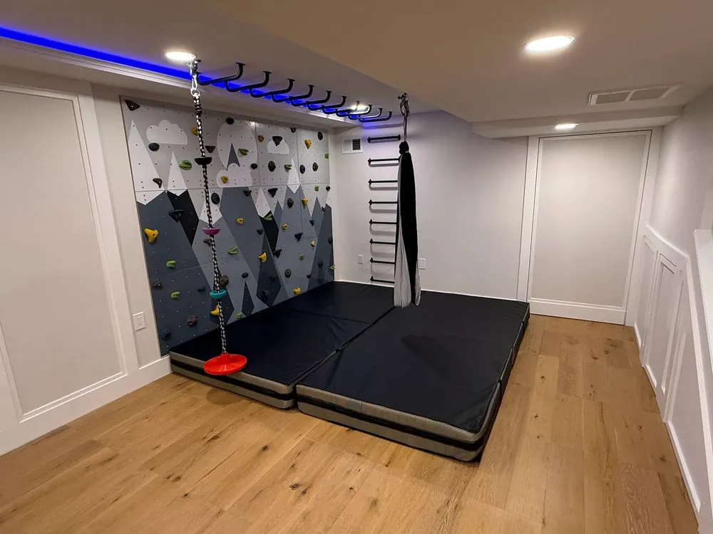 Basement play area with a mountain-painted climbing wall, ceiling monkey bars, swing, floor mats and white panel walls.