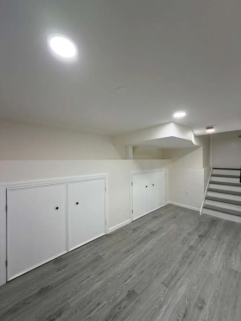 Finished basement with white knee-wall storage doors, gray vinyl plank floor and stairs with gray treads.