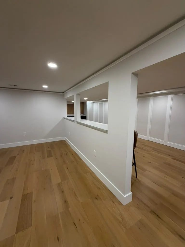Finished basement with wide oak plank floor, white walls, recessed lights and a half wall with a pass-through counter.