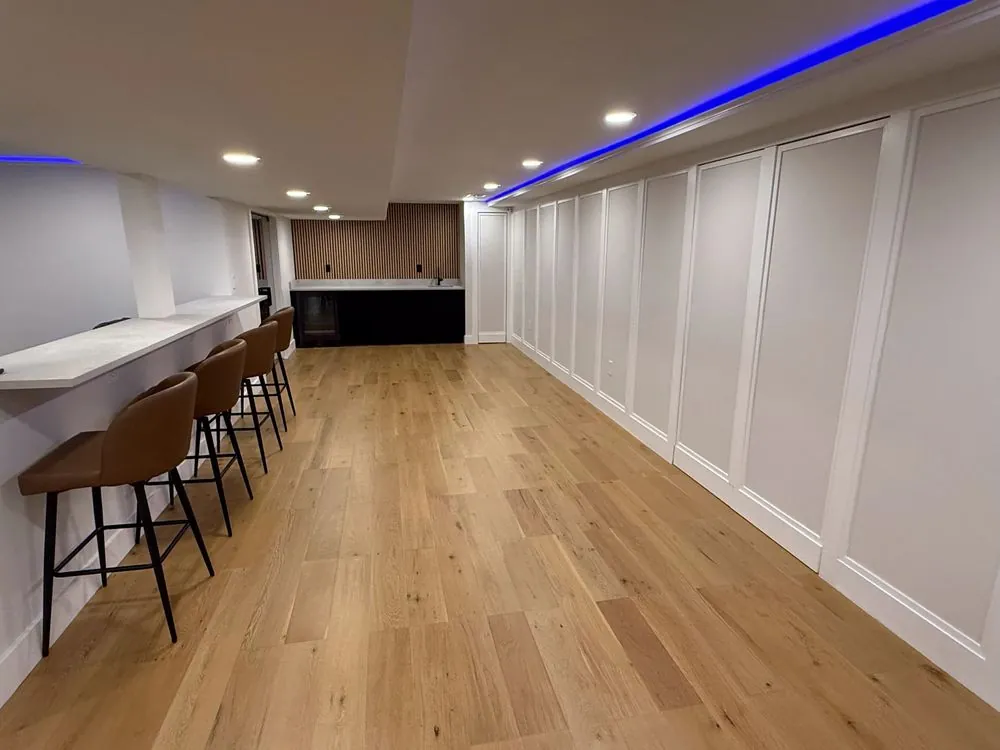 Finished basement with wide-plank oak floor, white panel wainscoting, a long counter with tan stools and a wet bar.