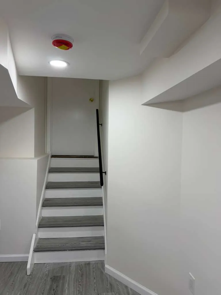 Finished basement stairway with gray treads, white risers, a black handrail and white walls with soffits.