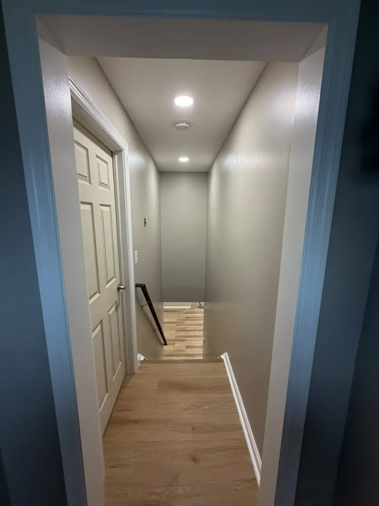 View down a basement stairway with gray walls, recessed lights, black handrail and light wood-look floor at the bottom.