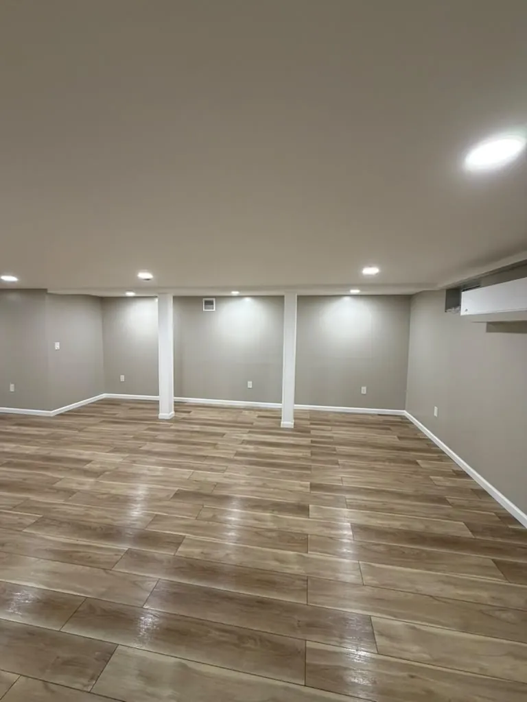 Open finished basement with wood-look vinyl plank floor, gray walls, white trim, wrapped columns and recessed lights.