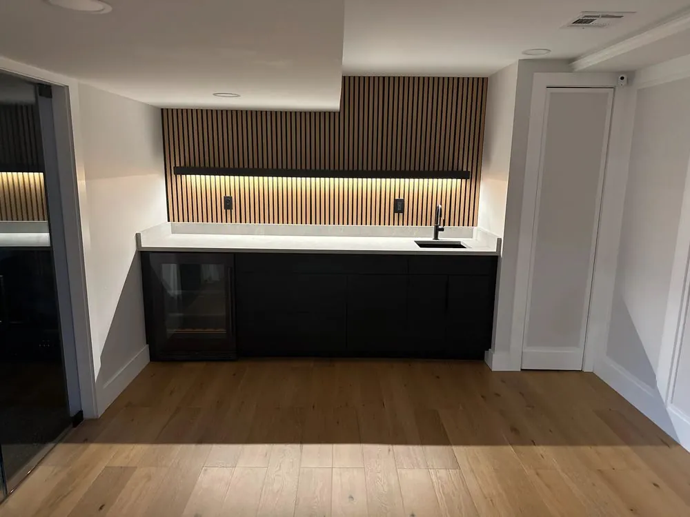 Basement wet bar with flat black cabinets, white quartz top, wine fridge, black faucet and a backlit wood slat wall.