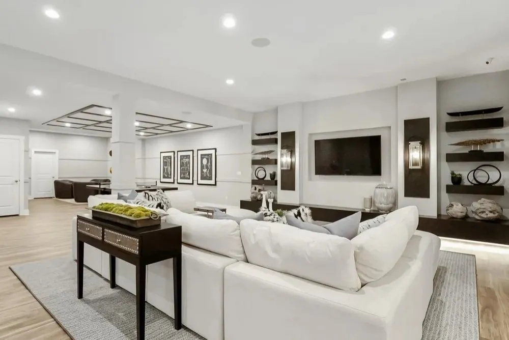 Large finished basement with white sectional sofa, TV wall with dark floating shelves, sconces and a coffered ceiling area.