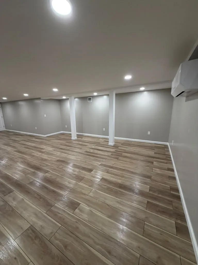 Wide finished basement with wood-look plank floor, white wrapped support columns, gray walls and a wall-mounted mini split.
