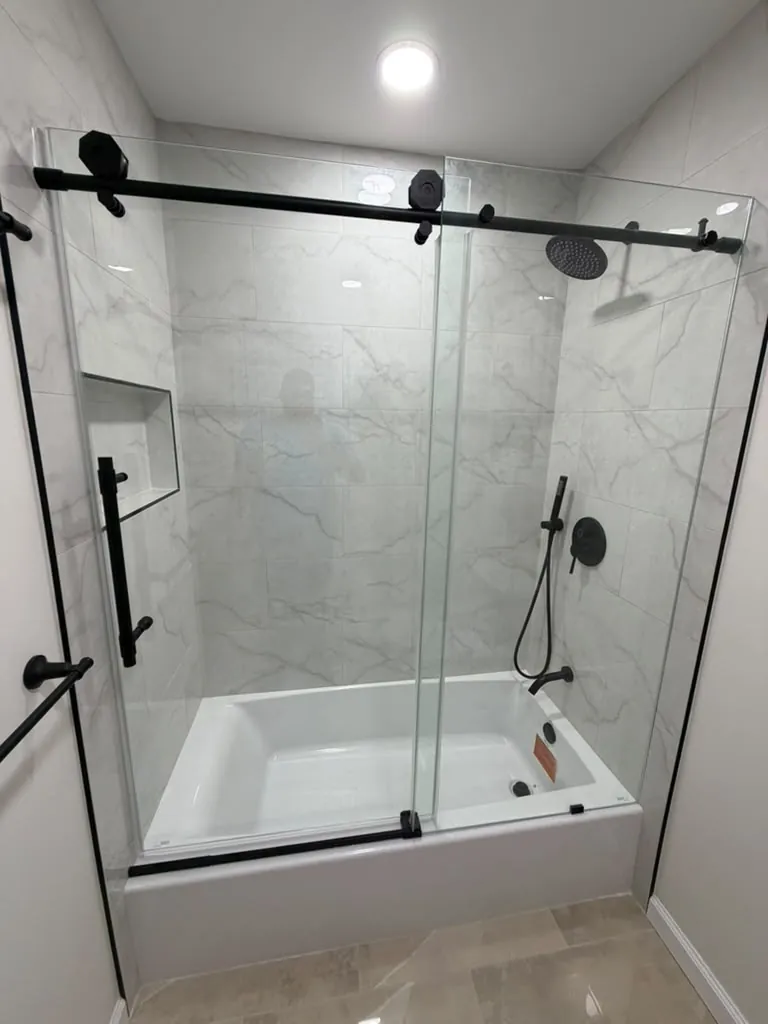 Alcove tub with white marble-look wall tile, a niche, matte black rain head, handheld and sliding glass door hardware.