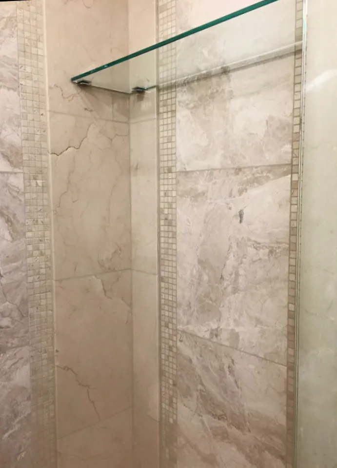 Shower wall with beige marble tile, vertical mosaic accent strips and a glass corner shelf.