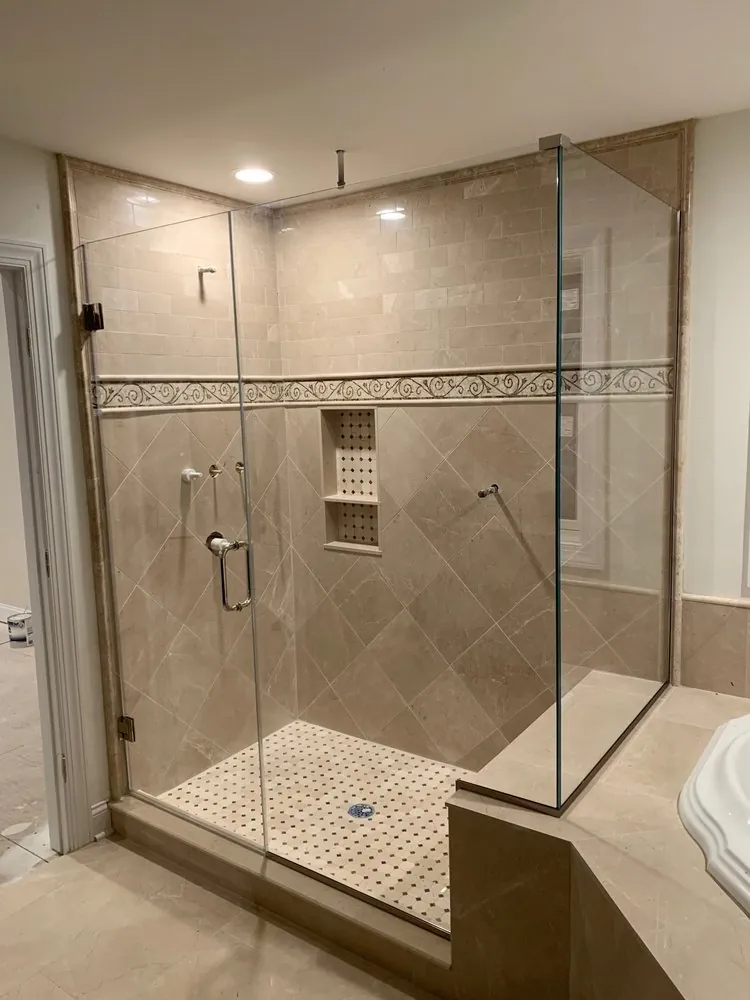 Frameless glass shower with beige marble tile set on the diagonal, scroll mosaic border, niche and dotted mosaic floor.