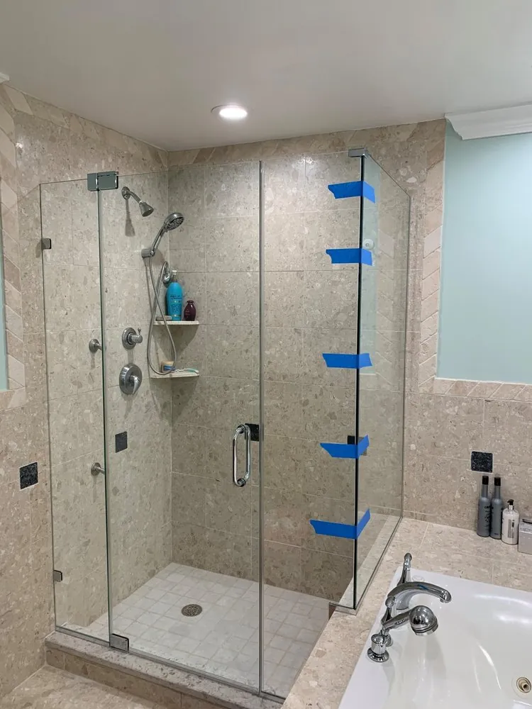 Frameless glass shower with beige speckled stone tile, chrome hand shower and corner shelves beside a jetted tub.