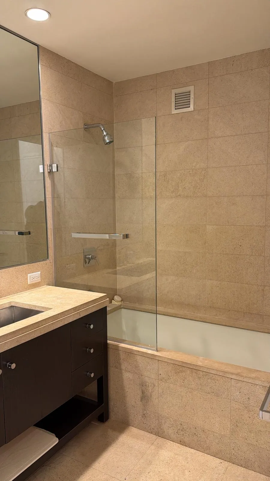 Bathroom with beige stone tile walls, soaking tub with fixed glass panel, chrome showerhead and dark wood vanity.