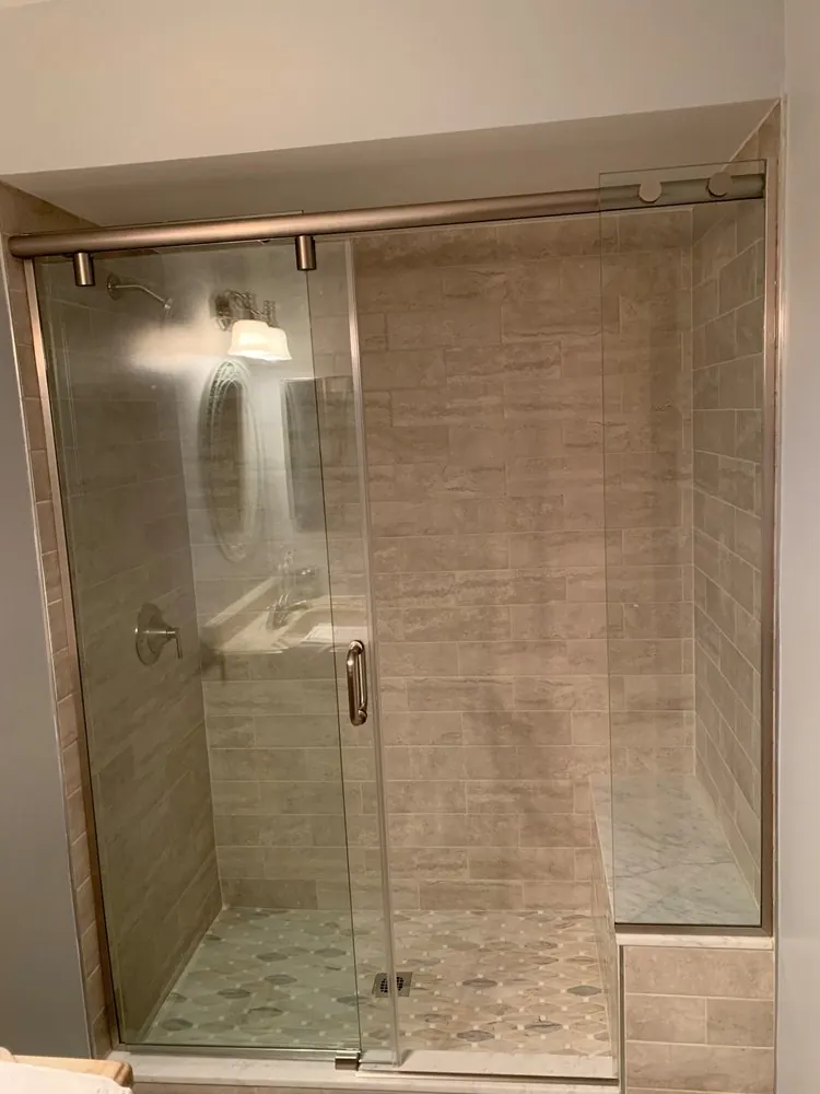 Shower with beige linear tile, marble-topped bench, mosaic floor and a sliding glass door on a brushed nickel rail.