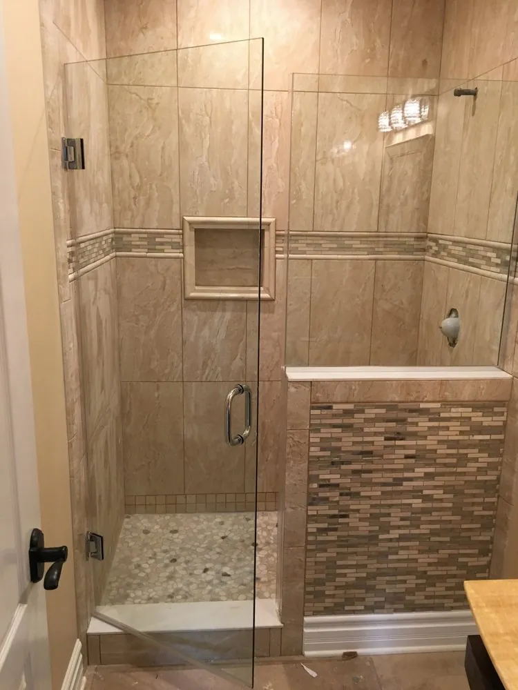 Glass shower with large beige tile, mosaic border band, framed niche, pebble floor and a mosaic-tiled half wall.