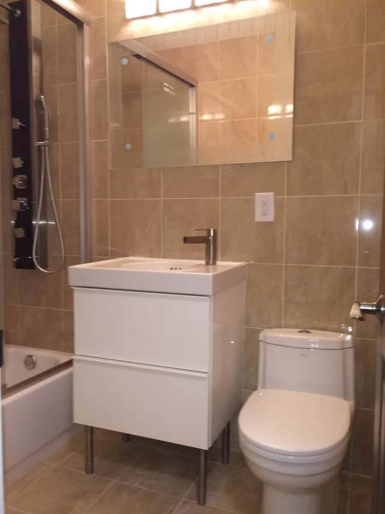 Bathroom with beige porcelain tile walls and floor, white two-drawer vanity on legs, mirror, toilet and shower panel.