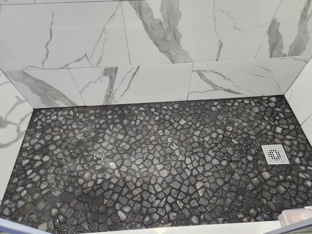 Close-up of a black pebble stone shower floor with square drain below white marble-look wall tile.