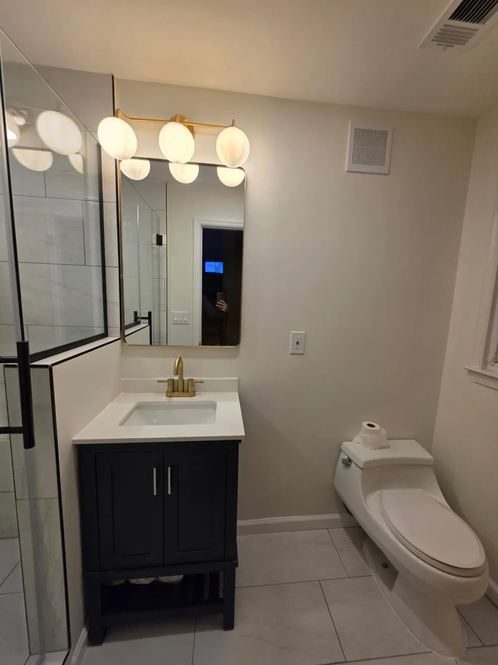 Black shaker vanity with white top and brass faucet under a gold-framed mirror and globe light, beside a toilet.