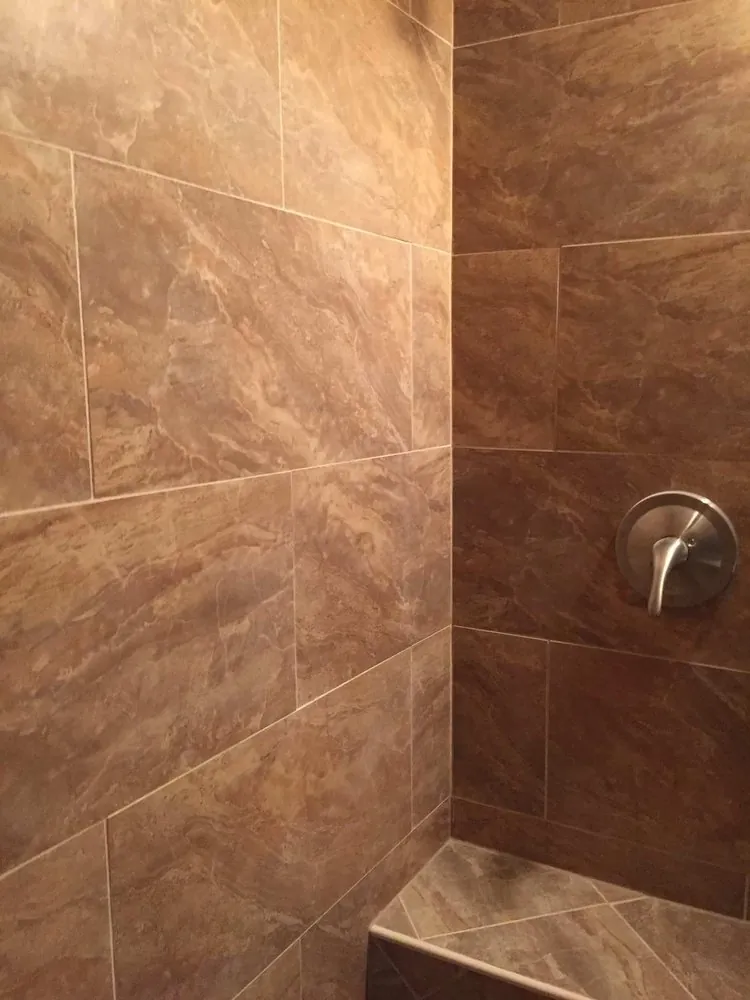 Shower corner with large brown marble-look porcelain tile, brushed nickel valve and a tiled corner bench.