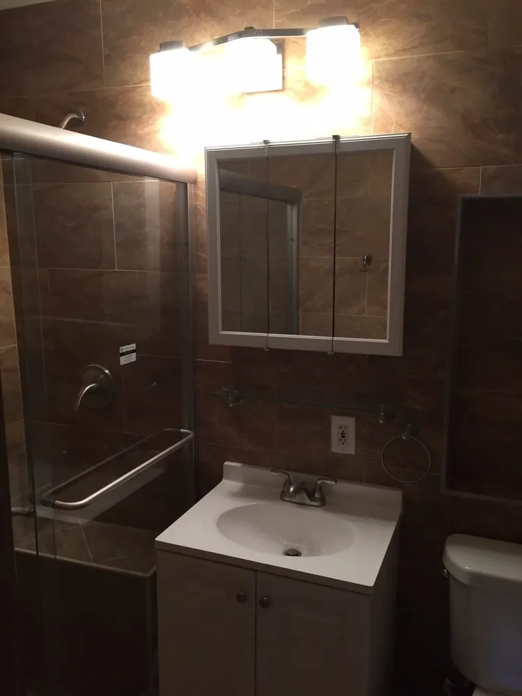 White vanity under a medicine cabinet and light bar, brown porcelain tile walls and a sliding glass shower door.