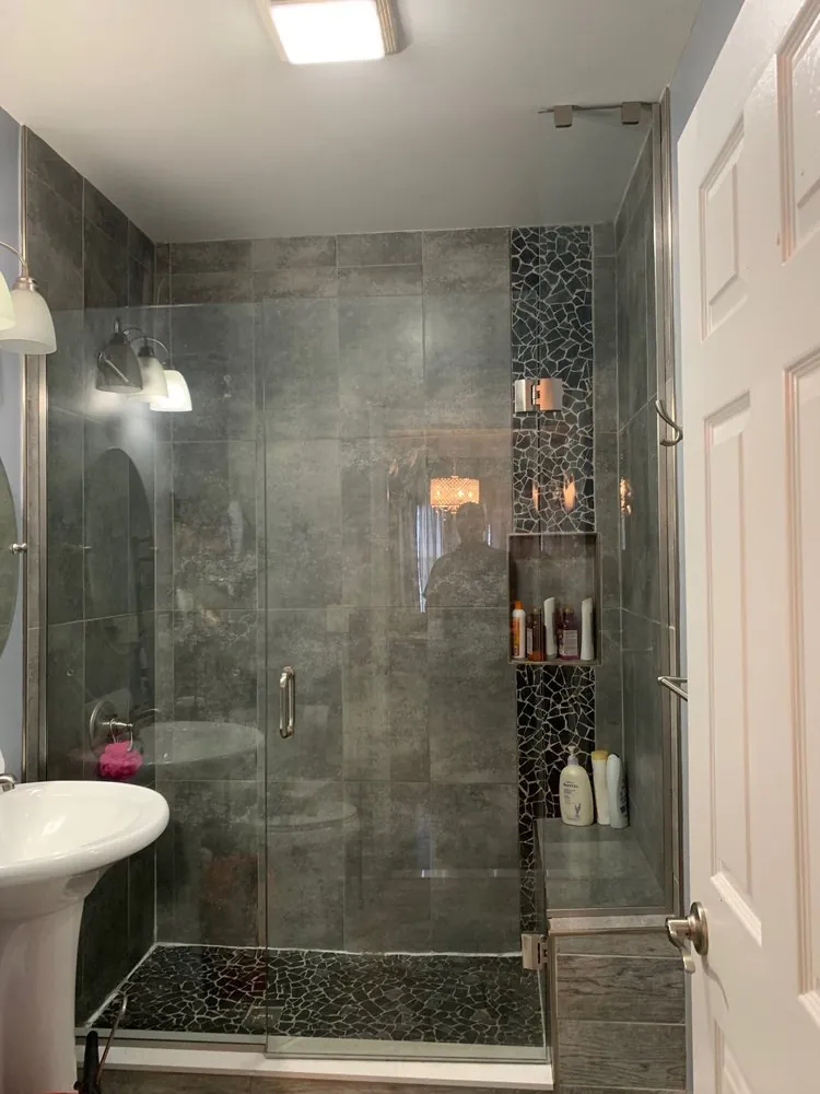 Glass shower with charcoal tile, black stone mosaic accent strip and niche, black pebble floor and white pedestal sink.