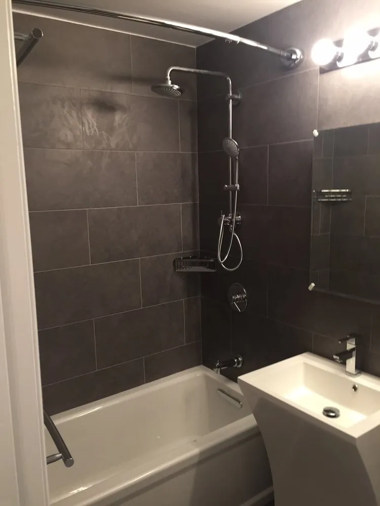 Bathtub with charcoal large-format tile surround, chrome shower column and rain head, and a white pedestal sink.