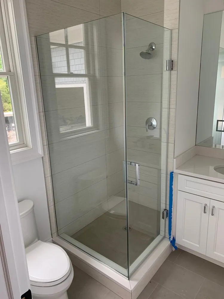 Frameless glass corner shower with beige linen-look wall tile, chrome fixtures, next to a white vanity and toilet.