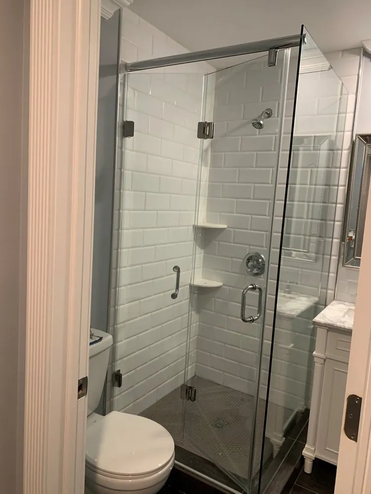 Frameless corner glass shower with white beveled subway tile, corner shelves and gray hex floor next to a white vanity.