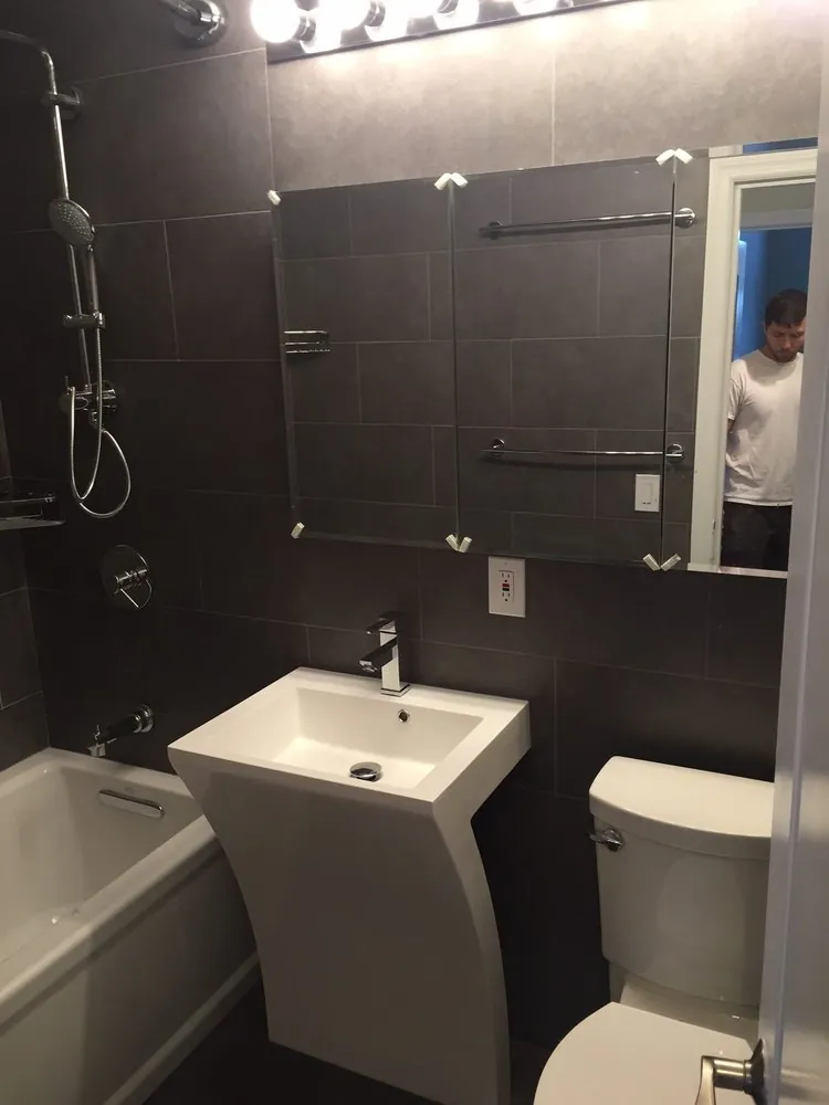 Dark gray tiled bathroom with a modern white pedestal sink, mirror, toilet and tub with handheld shower.