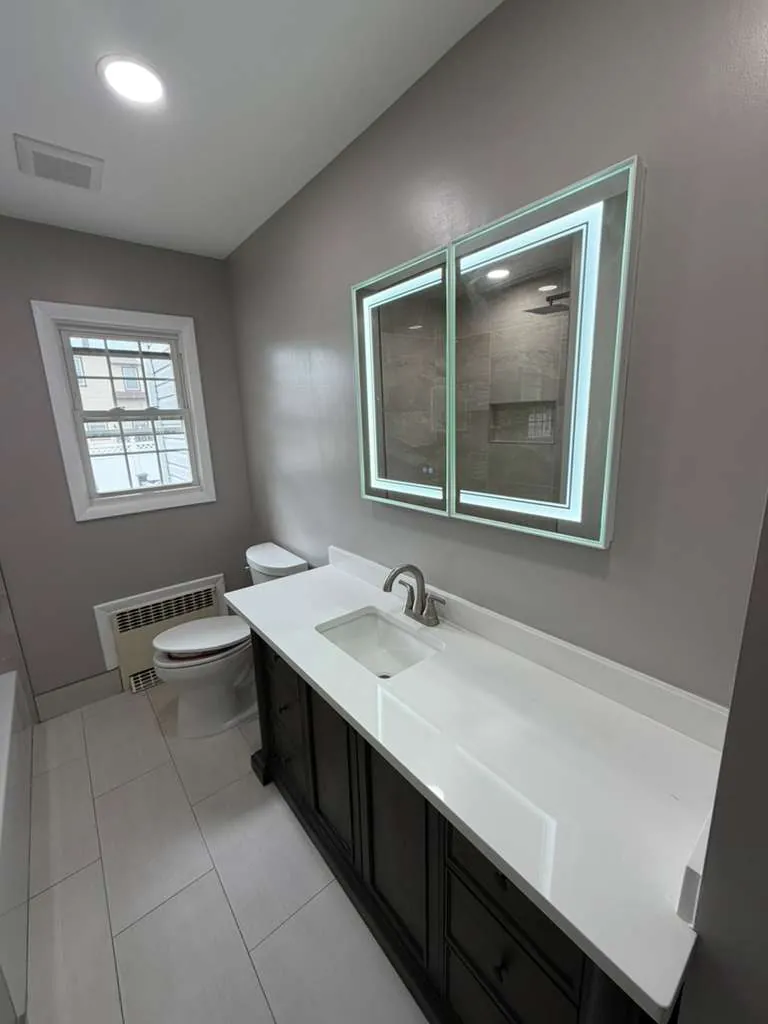 Bathroom with a long dark wood vanity, white top, undermount sink, lighted mirror cabinet and light gray floor tile.