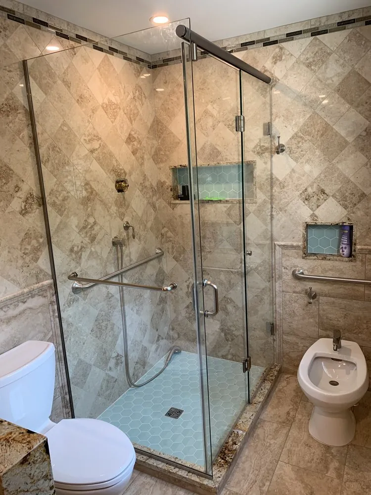 Glass shower with diagonal beige marble tile, aqua hexagon floor, two niches and grab bars, beside a toilet and bidet.