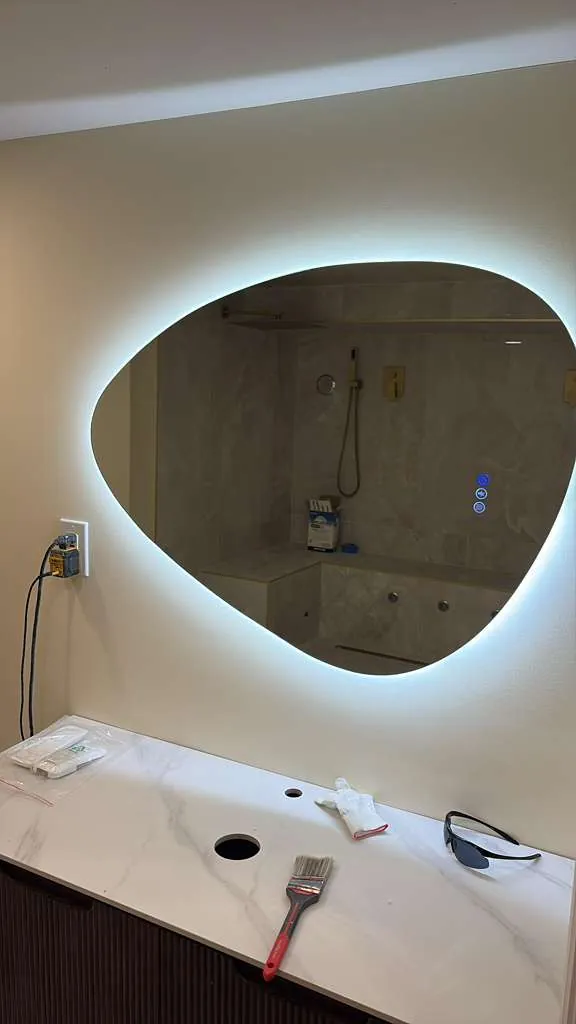 Organic-shaped backlit mirror above a white marble-look vanity top with no sink yet, paintbrush and tools on the counter.