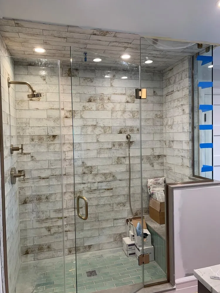 Large glass shower with brass fixtures, distressed white plank tile on walls and ceiling, green glass floor, supplies inside.
