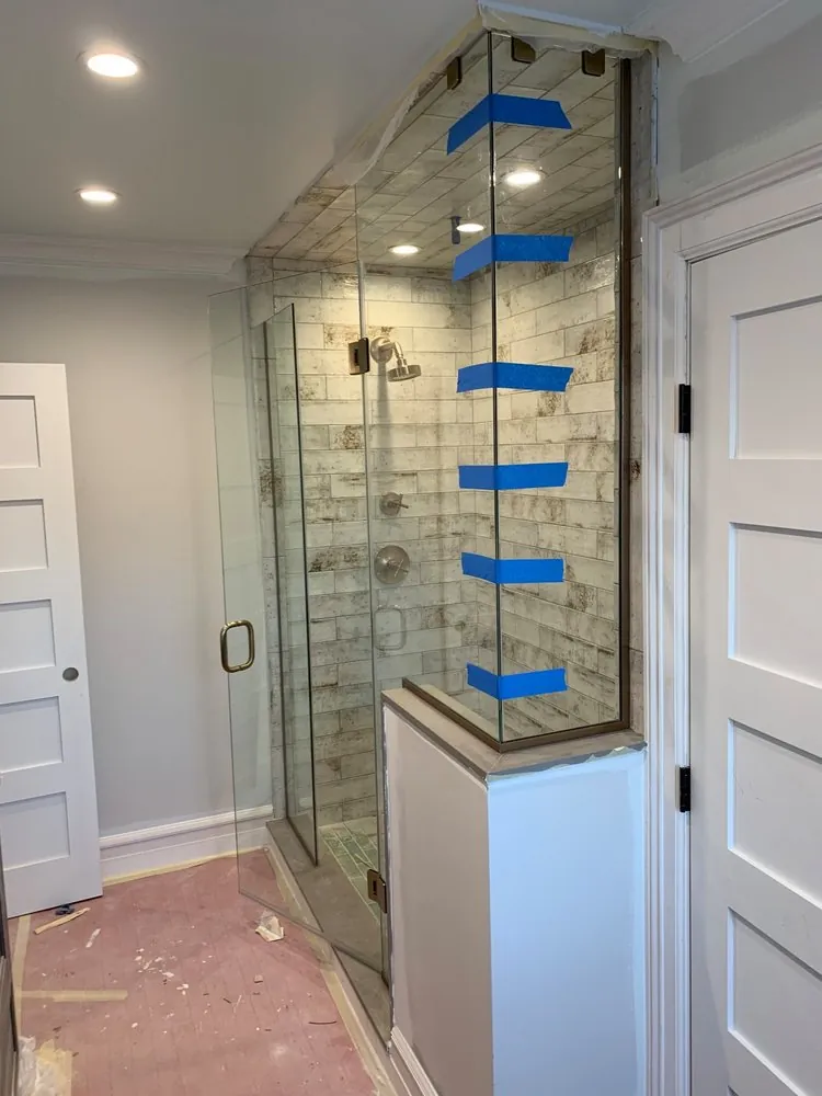 Frameless glass shower with antique white subway tile and bronze hardware, glass taped, floor still covered.