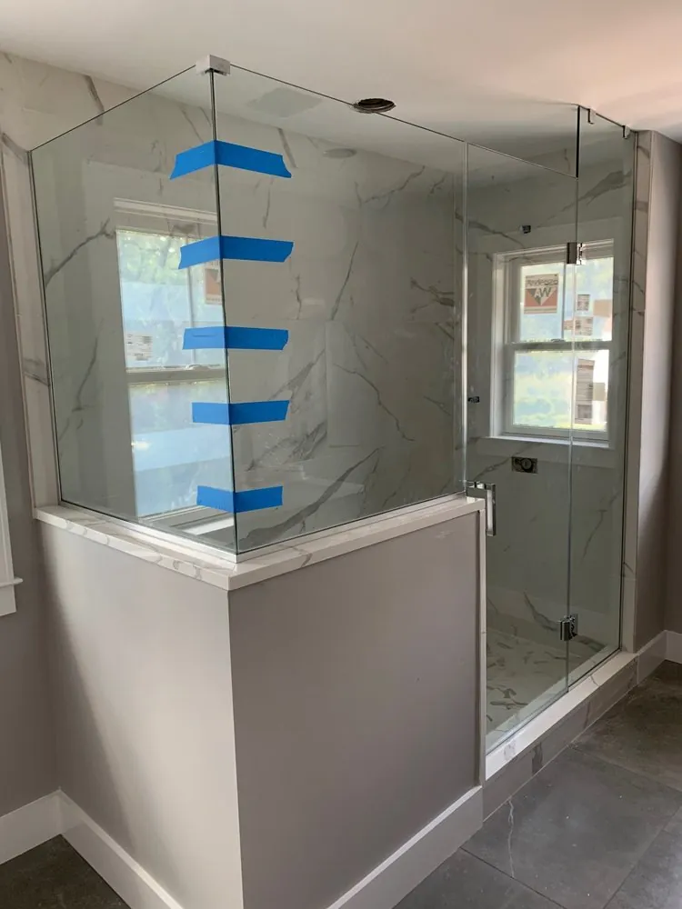 Glass shower enclosure on a gray knee wall with white marble-look porcelain walls and dark tile floor, new windows.