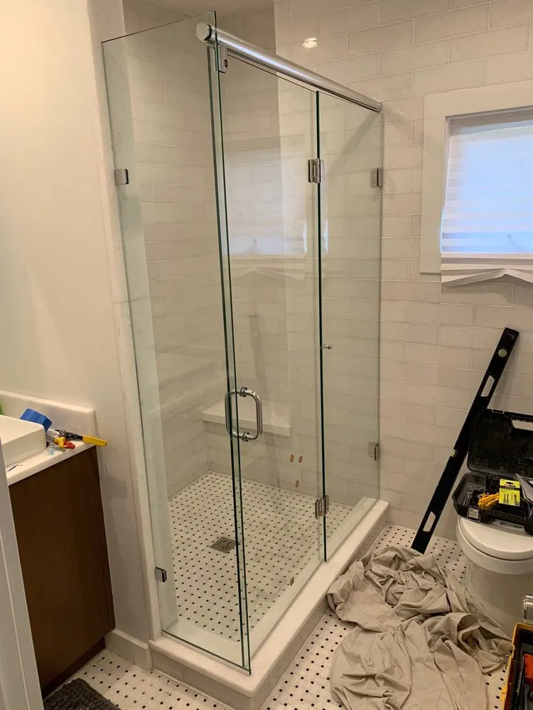 Frameless glass shower being installed with white subway tile and black dot basketweave floor, tools nearby.